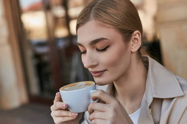 Charmante blonde Frau im Trenchcoat genießt ihre Tasse Cappuccino im Café, entspannte Atmosphäre — Stockfoto