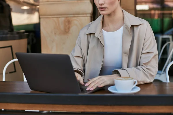 Cropped view of young woman in trench coat using laptop near cup of cappuccino in cafe, remote work — Stock Photo