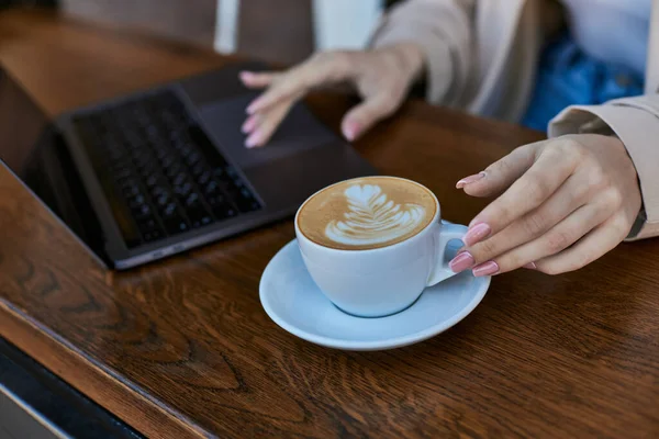 Cropped shot of young woman in trench coat using laptop near cup of cappuccino in cafe, remote work — Stock Photo