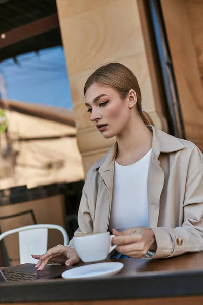 Blonde young woman in trench coat using laptop near cup of cappuccino in cafe, remote work — Stock Photo