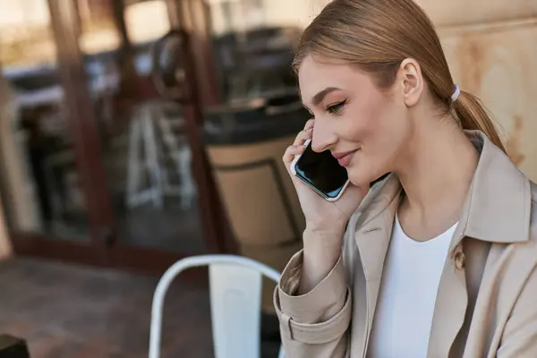 Счастливая молодая женщина в осеннем бежевом плаще улыбается и разговаривает по смартфону в кафе — стоковое фото