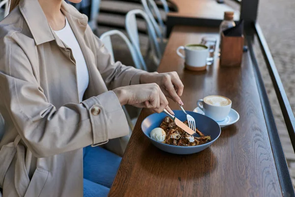 Cropped view of woman in trench coat eating belgian waffles with ice cream next to cup of coffee — Stock Photo
