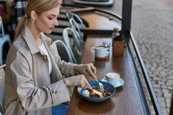 Rubia joven en gabardina comiendo sus gofres belgas con helado al lado de una taza de café - foto de stock