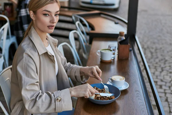 Hübsche junge Frau im Trenchcoat isst ihre belgischen Waffeln mit Eis neben einer Tasse Kaffee — Stockfoto