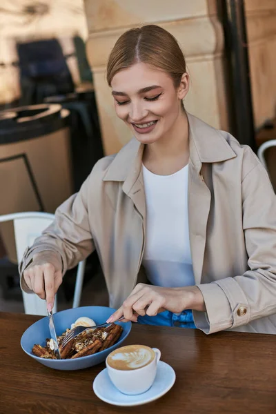 Happy young woman in trench coat eating her belgian waffles with ice cream next to cup of coffee — Stock Photo