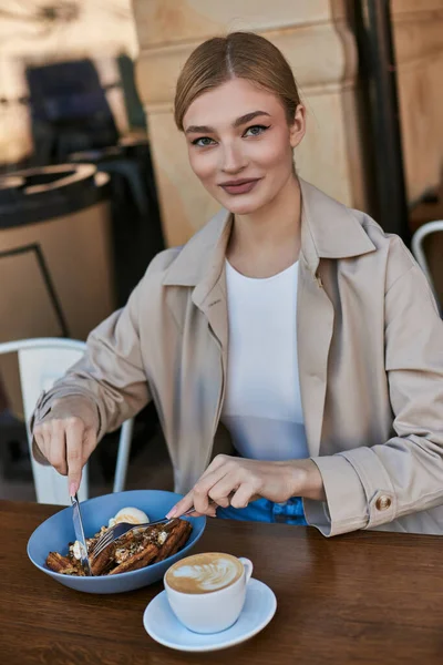 Happy young woman in trench coat enjoying her belgian waffles with ice cream next to cup of coffee — Stock Photo