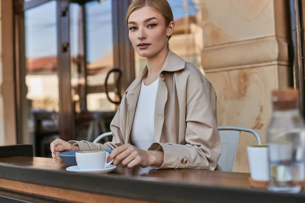 Schöne junge Frau in stylischem Trenchcoat sitzt neben Teller und Tasse Kaffee im modernen Café — Stockfoto