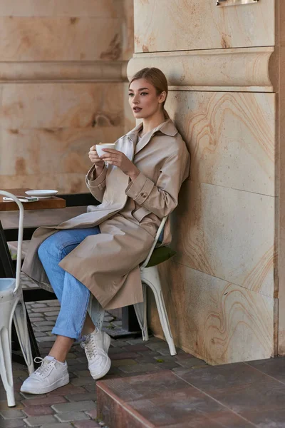 Beautiful young woman in stylish trench coat sitting and holding cup of coffee inside of modern cafe — Stock Photo