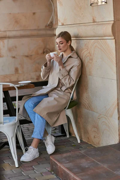 Приваблива молода жінка в стильному тренч пальто сидить і тримає чашку кави всередині кафе — стокове фото