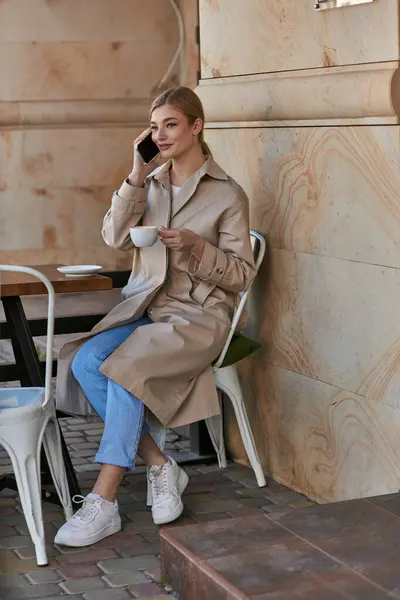 Щаслива молода жінка в стильному тренч пальто розмовляє на смартфоні і тримає чашку кави в кафе — стокове фото