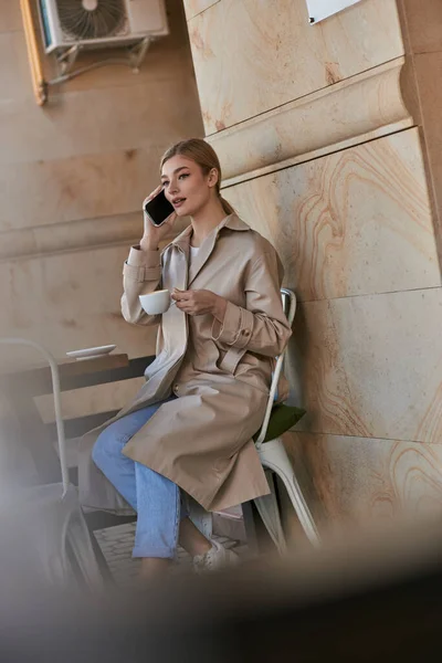 Hübsche junge Frau im schicken Trenchcoat, die mit dem Smartphone spricht und im Café eine Tasse Kaffee hält — Stockfoto