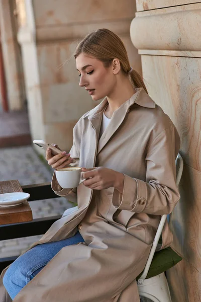 Attrayant jeune femme dans élégant trench coat en utilisant smartphone près de tasse de café sur la table dans le café — Photo de stock
