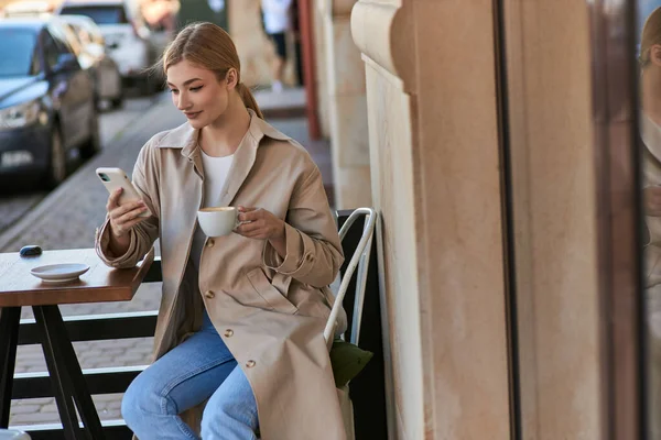 Привлекательная женщина в стильном плаще с помощью смартфона возле чашки кофе на столе в кафе — стоковое фото
