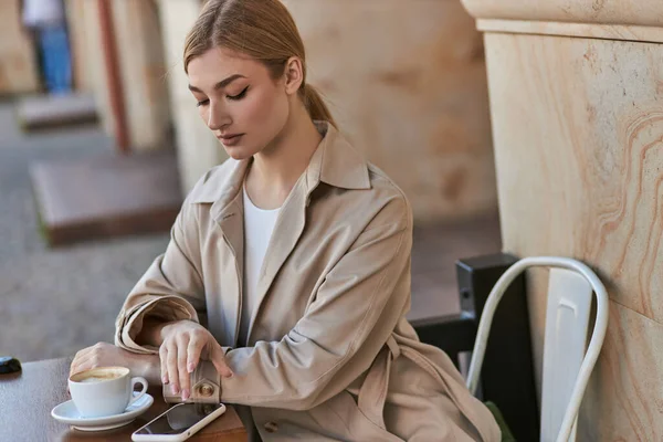 Blonde young woman in stylish trench coat using smartphone near cup of coffee on table in cafe — Stock Photo