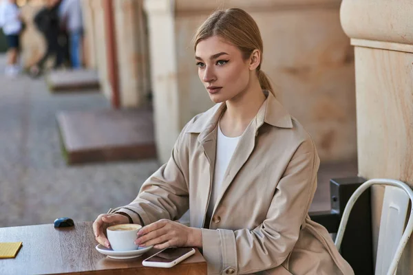 Bionda giovane donna in trench seduta vicino a tazza di caffè e smartphone sul tavolo in caffè — Foto stock