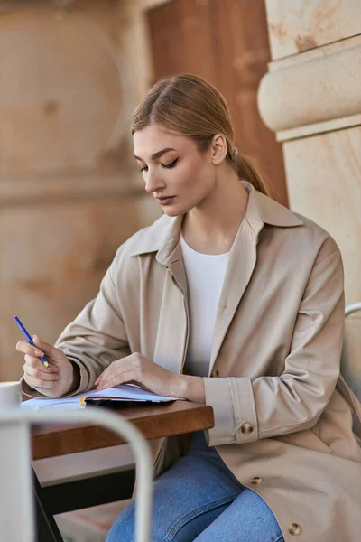 Pretty young woman in trench coat taking notes in notebook near cup of coffee in cafe, planning day — Stock Photo