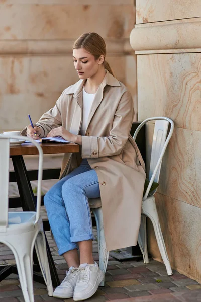 Blonde young woman in trench coat taking notes in notebook near cup of coffee in cafe, planning day — Stock Photo