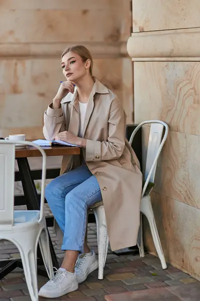 Thoughtful blonde woman in trench coat sitting at table and holding pen near chin, writing diary — Stock Photo