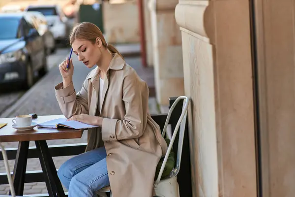 Nachdenkliche Frau im Trenchcoat sitzt mit Kaffeetasse am Tisch und hält Stift in der Hand, schreibt Tagebuch — Stockfoto