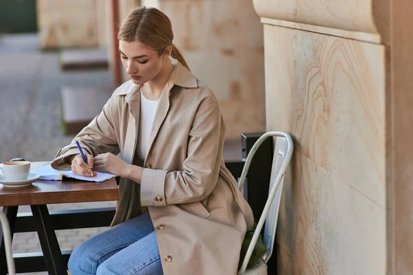 Nachdenkliche junge Frau in schickem Trenchcoat, in Notizbuch neben Kaffeetasse, Tagebuch — Stockfoto