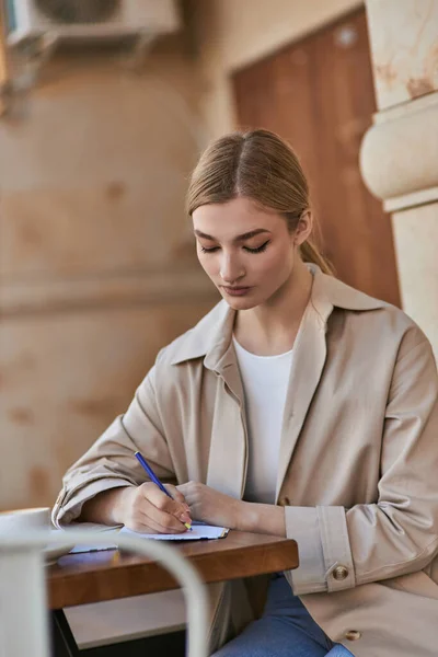 Pretty young woman in stylish trench coat writing in notebook near cup of coffee, taking notes — Stock Photo