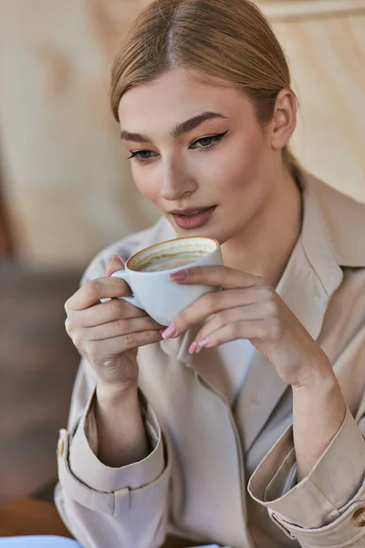 Elegant blonde woman in stylish trench coat holding cup with coffee while sitting in cafe, beverage — Stock Photo