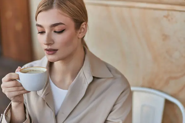 Élégante femme blonde dans un élégant trench coat tenant une tasse de café tout en étant assis dans un café, cappuccino — Photo de stock