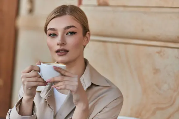 Élégante jeune femme dans un élégant trench coat tenant tasse de café tout en étant assis dans un café, boisson — Photo de stock