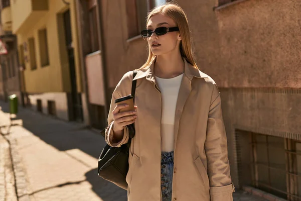 Красива жінка в стильному тренч пальто тримає винос кави, стоячи на вулиці в сонячний день — стокове фото