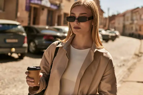 Blonde femme dans élégant trench coat tenant café pour aller tout en se tenant debout dans la rue dans la journée ensoleillée — Photo de stock