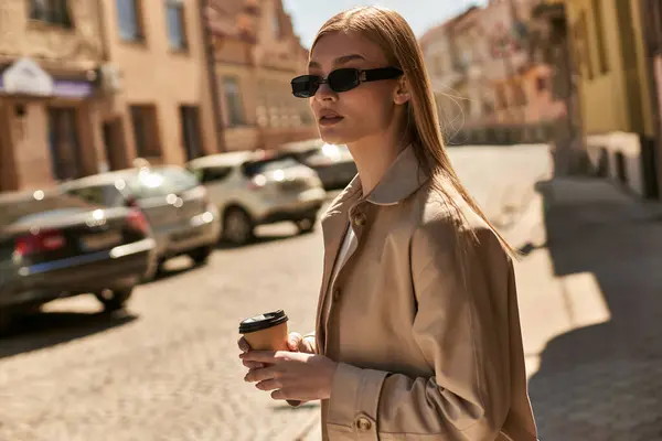 Блондинка в стильному тренч пальто тримає каву, щоб піти під час прогулянки на вулиці в сонячний день — стокове фото