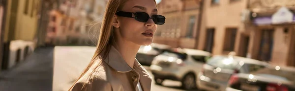 Jeune femme blonde dans un élégant trench coat et lunettes de soleil marchant dans la rue par temps ensoleillé, bannière — Photo de stock