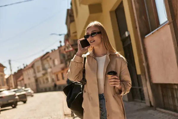 Mujer rubia en gabardina y gafas de sol hablando en smartphone y sosteniendo café para salir a la calle - foto de stock