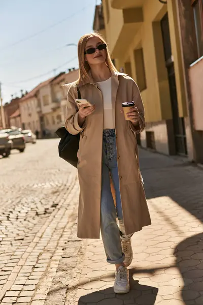 Joyeuse jeune femme en trench coat et lunettes de soleil tenant smartphone et café pour aller dans la rue — Photo de stock