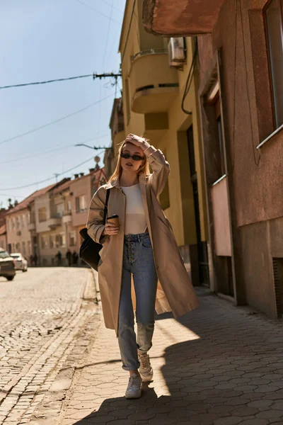 Elegante giovane donna in trench e occhiali da sole che tengono il caffè per andare e camminare per strada — Foto stock