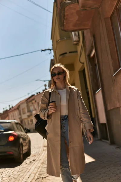 Блондинка в плаще и солнцезащитных очках держа кофе идти и ходить по улице — стоковое фото