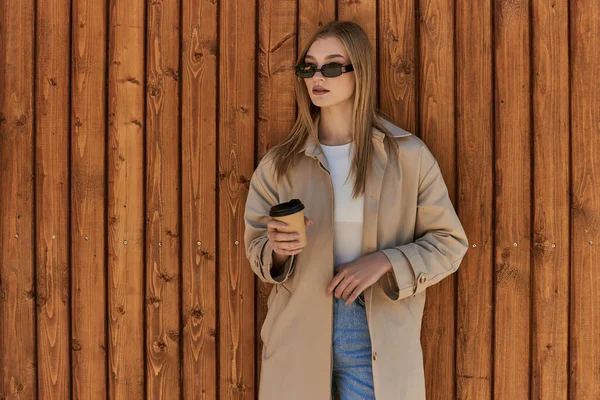 Blonde jeune femme en trench coat et lunettes de soleil tenant café pour aller et debout près de clôture en bois — Photo de stock
