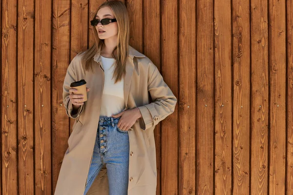 Блондинка в стильному траншеї пальто і сонцезахисні окуляри тримають каву, щоб піти і стояти біля паркану — стокове фото