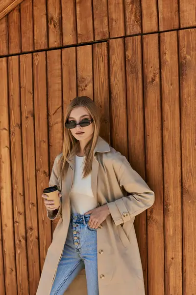 Blondine in Trenchcoat und Sonnenbrille hält Coffee to go in der Hand und steht mit der Hand in der Tasche — Stockfoto