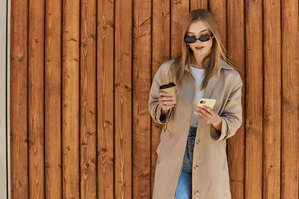 Блондинка в плаще и солнцезащитных очках держит кофе, чтобы пойти и использовать смартфон рядом с забором — стоковое фото