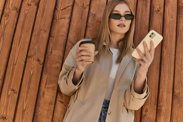 Young woman in trench coat and sunglasses holding coffee to go and using smartphone near fence — Stock Photo