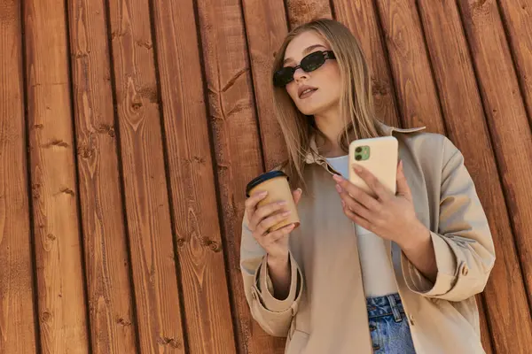 Femme élégante en trench coat et lunettes de soleil tenant café pour aller et en utilisant smartphone près de la clôture — Photo de stock