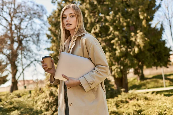 Young blonde woman in trench coat walking with laptop and takeaway coffee, freelancer in city — Stock Photo