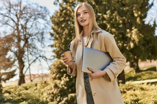 Happy blonde woman in trench coat walking with laptop and takeaway coffee, freelancer in city — Stock Photo