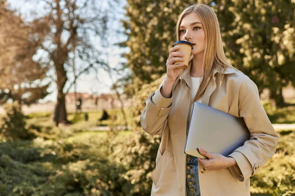 Jeune femme blonde en trench coat tenant ordinateur portable et boire du café à emporter, pigiste en ville — Photo de stock