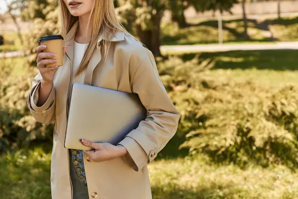 Обрізана молода жінка в траншеї пальто тримає ноутбук і виймає каву, фрілансер в місті — стокове фото