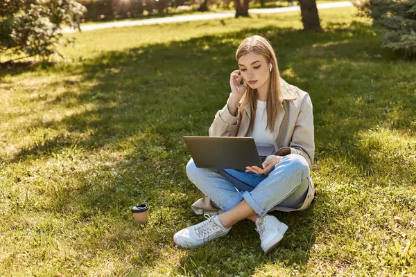 Mujer rubia joven en auriculares y gabardina sentado en la hierba cerca de la taza de papel y el uso de ordenador portátil - foto de stock