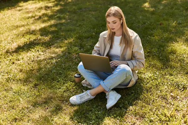 Blonde woman in earphones and trench coat sitting on grass near paper cup and using laptop — Stock Photo