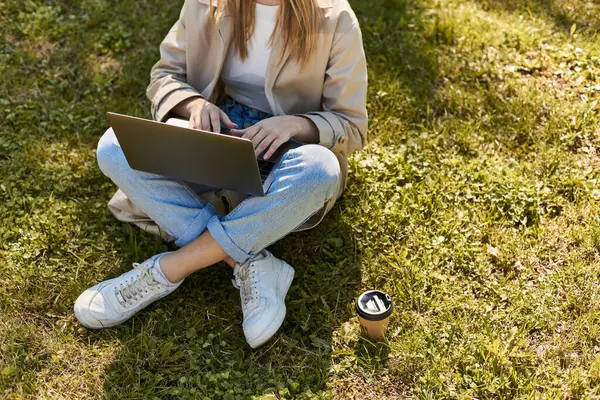 Cropped woman in jeans and trench coat sitting on green lawn near paper cup and using laptop — Stock Photo