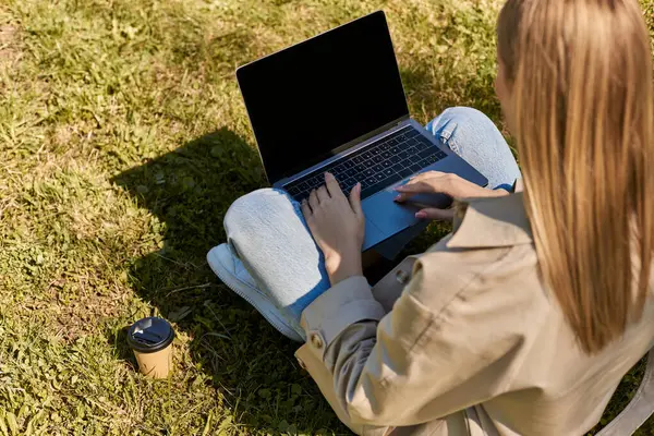 Overhead view, woman in jeans and trench coat sitting on green lawn near paper cup and using laptop — Stock Photo